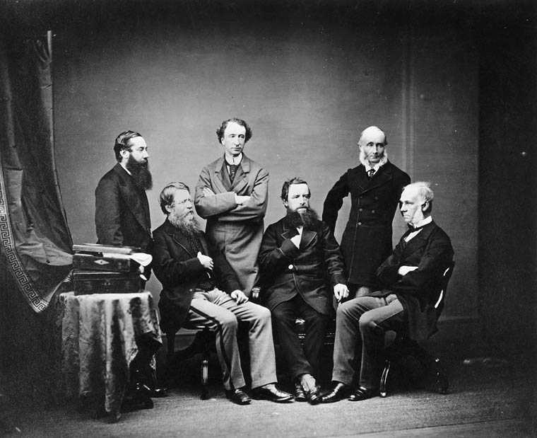 Historical black and white photography of six men in dress coats, three standing, three seated, grouping for the picture.