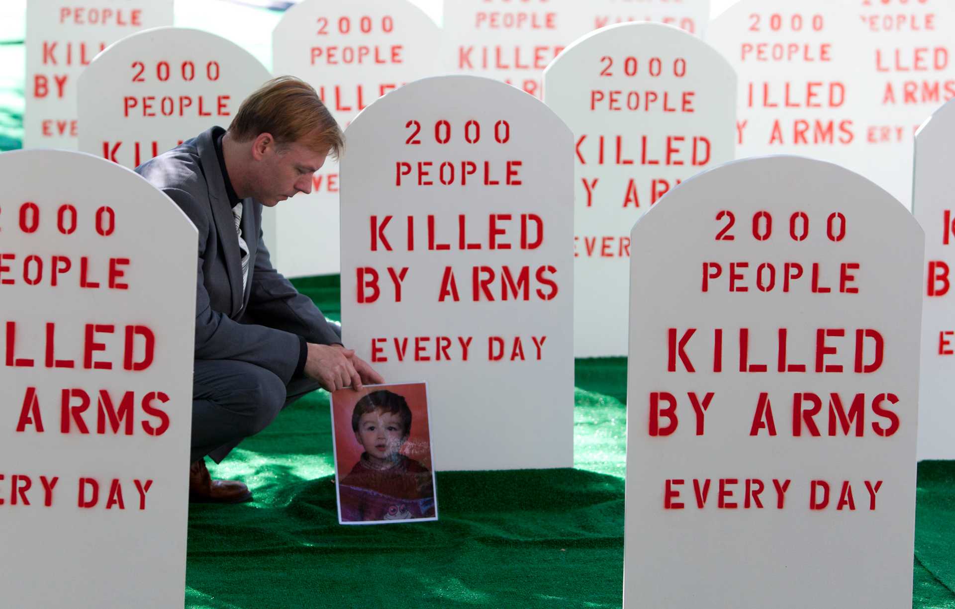 Middle-aged in a grey suit kneels among tombstone-shaped placards reading: 2000 people killed by arms every day. He is laying a large photograph of a young boy.